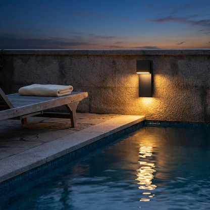 Honfleur black outdoor wall light glowing on a poolside wall at dusk